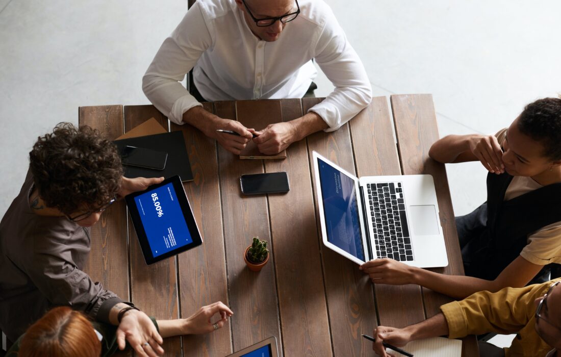 Visão superior de mesa de trabalho com dois homens e três mulheres, usando tablet e notebook, em espaço de integração para estágio.