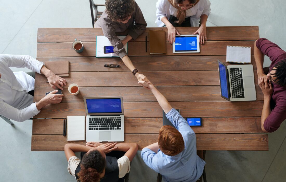 Imagem de vista superior mostrando jovens em mesa de trabalho com computadores e tablets, ilustrando texto sobre vagas de estágio e trainee disponíveis para alunos da PUC Minas